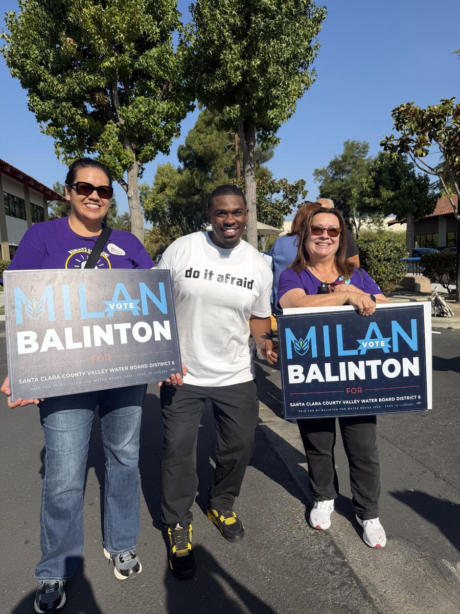 Milan Balinton stands between two supporters holding campaign signs in a parking lot