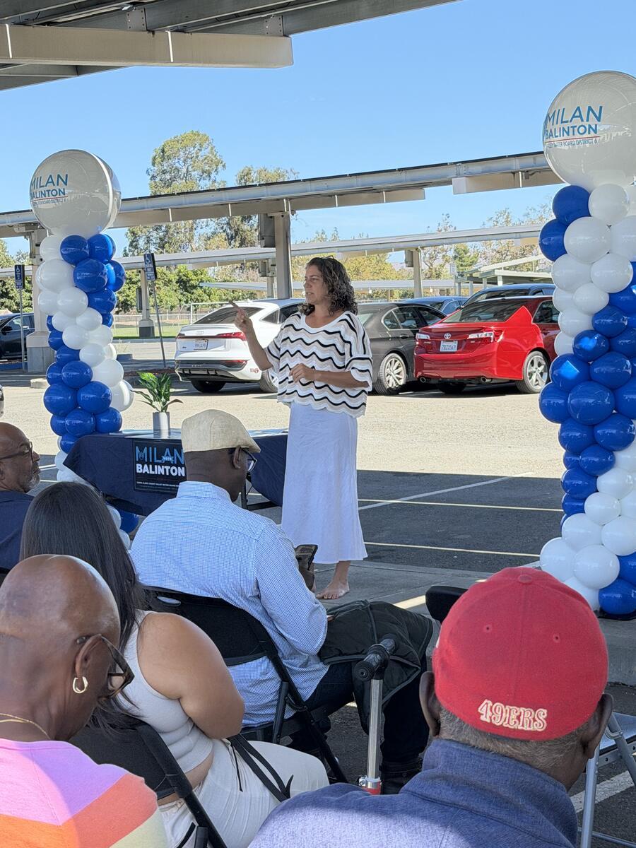 A speaker addresses a seated crowd between blue and white balloon arches at a campaign rally
