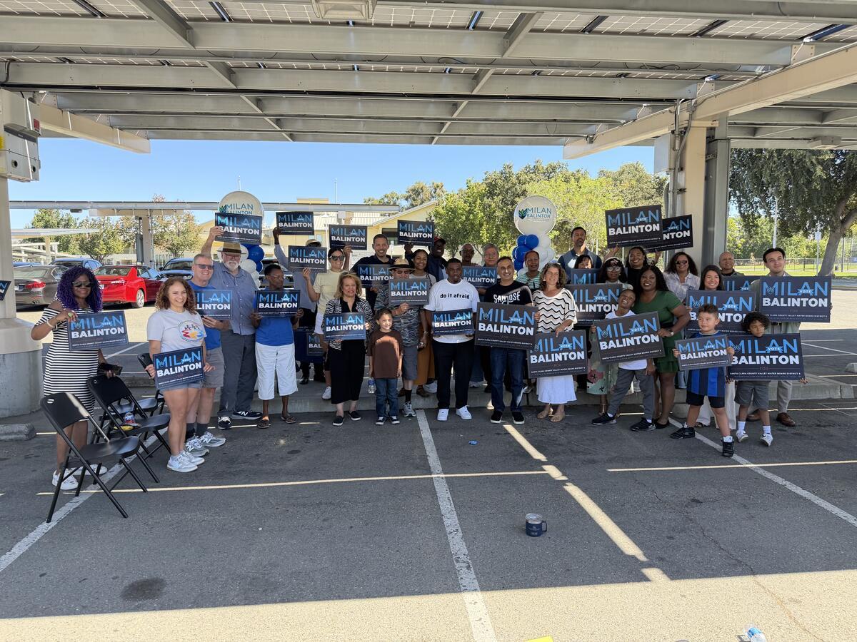A crowd of supporters holds Milan Balinton signs under a solar canopy