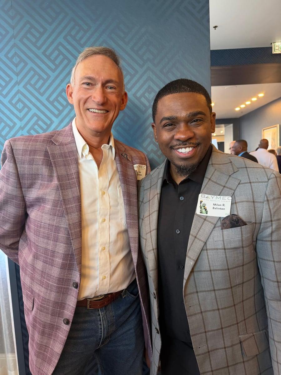 An event guest in a checked jacket stands with Milan Balinton at a BAYMEC gathering