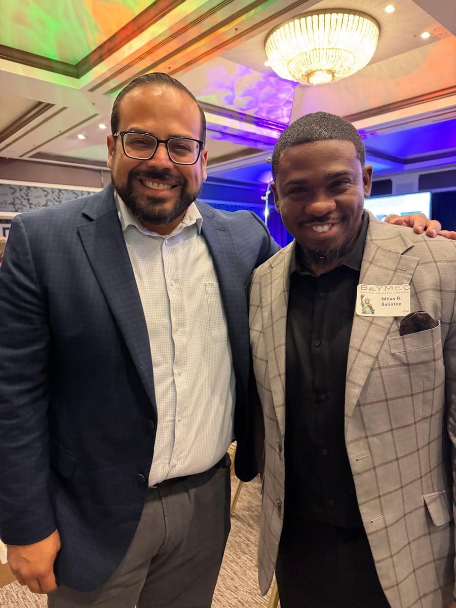An event guest wearing glasses and a navy suit stands with Milan Balinton at a BAYMEC gathering