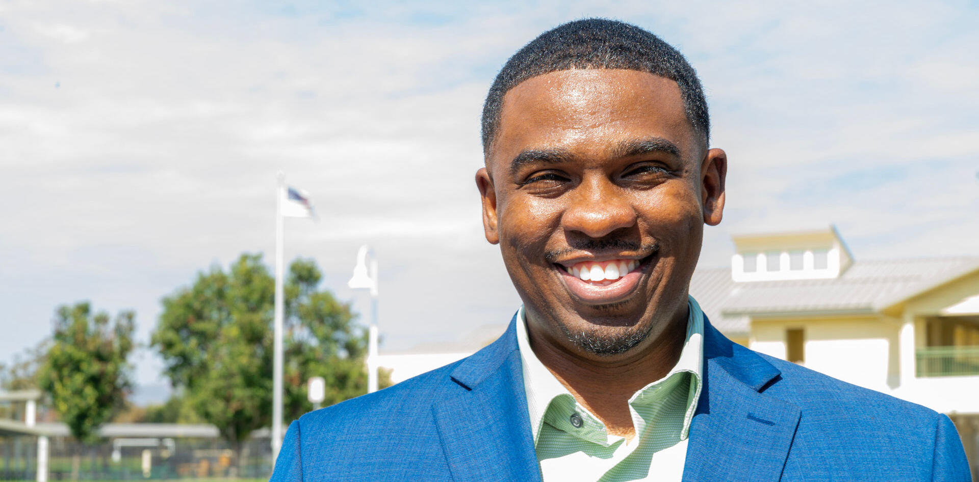 Mr. Milan Balinton, smiling for a headshot in landscape. Wearing a brilliant blue blazer with a pastel light green shirt, open button, no tie.